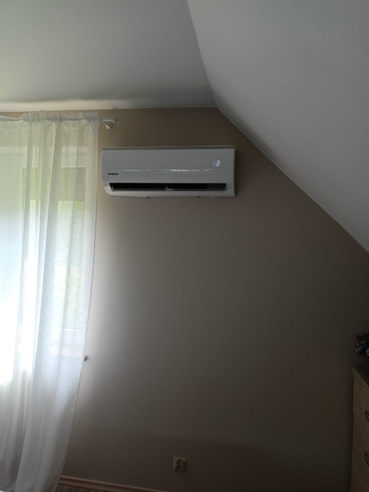 Zamontowany klimatyzator Kaisai na ścianie pokoju na poddaszu, obok okna z białymi zasłonami. Widoczny wyświetlacz z temperaturą 22 stopnie.