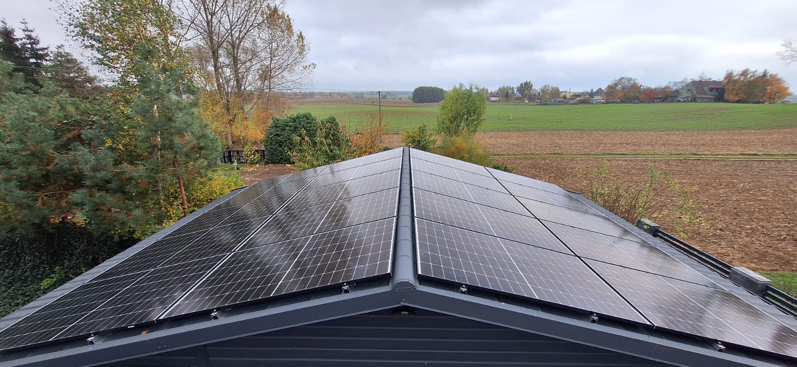 Panele fotowoltaiczne na dachu budynku z szarej blachy, widok z góry, w tle jesienny krajobraz pól i drzew pod pochmurnym niebem.