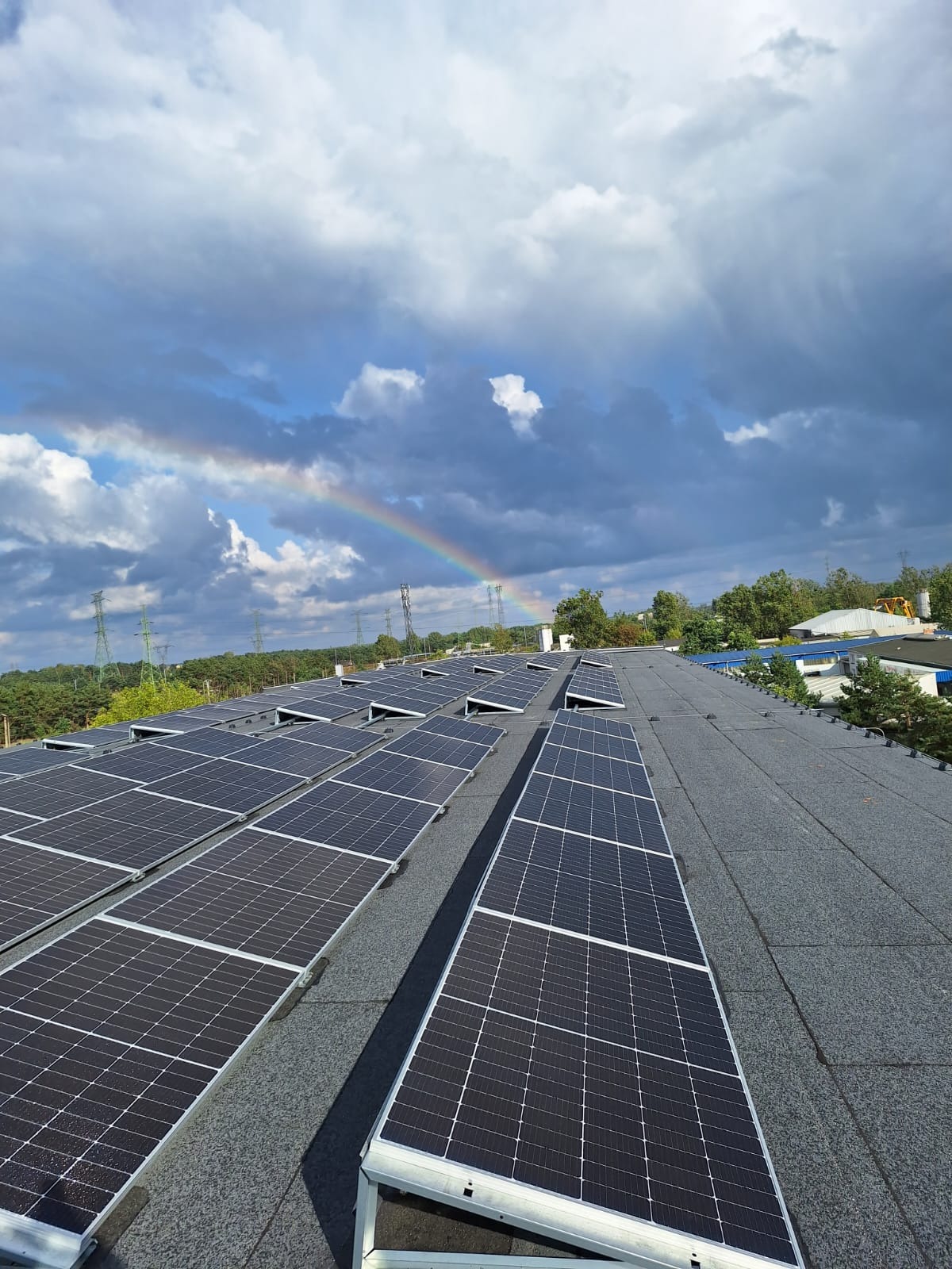 Instalacja fotowoltaiczna na dachu budynku, widok z góry, z tęczą na tle pochmurnego nieba. Panele w srebrnych ramach ułożone rzędami.