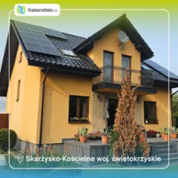 Żółty dom jednorodzinny z panelami fotowoltaicznymi na dachu, ozdobnym balkonem i kamienną elewacją. Widoczne kwiaty w donicach i zadbany ogród.