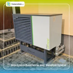 Szara pompa ciepła Calla Verde na zewnątrz budynku, widoczne rury i fragment elewacji. Ujęcie z bliska, z niskiej perspektywy.