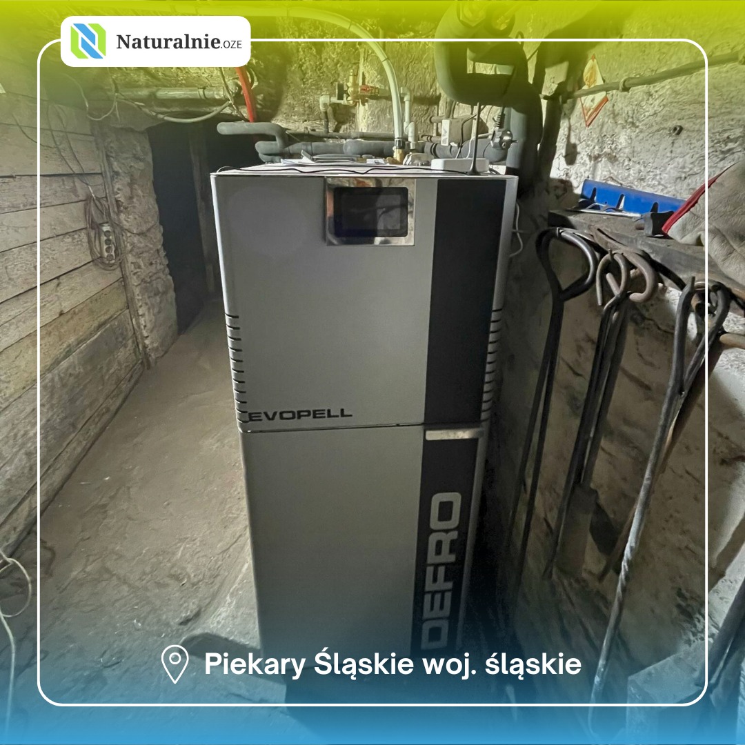 Szary piec Defro Evopell w piwnicy z kamienną ścianą i drewnianymi drzwiami. Obok pieca wiszą metalowe narzędzia. Ujęcie z dołu.