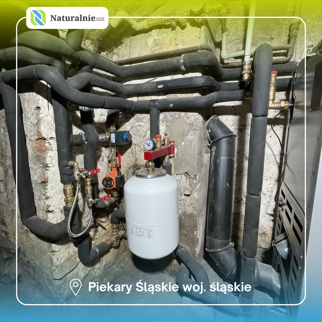 Instalacja grzewcza z czarnymi rurami i białym zbiornikiem wyrównawczym. Realizacja w piwnicy w Szydłowcu. Widoczne liczne zawory i manometry.