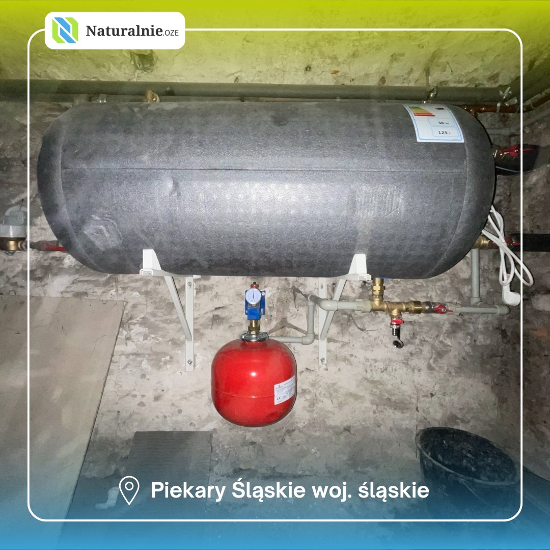 Szary zbiornik akumulacyjny ciepła z czerwonym naczyniem wzbiorczym, zamontowany na ścianie z widocznymi rurami i armaturą w piwnicy.