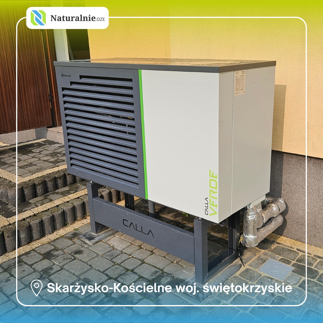 Szara pompa ciepła Calla Verde na zewnątrz budynku, widoczne rury i fragment elewacji. Ujęcie z bliska, z niskiej perspektywy.