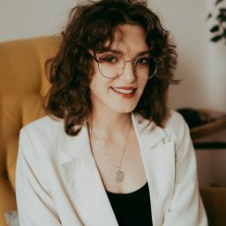 Poradnia psychoterapii MIND-TALK-THERAPY - Uśmiechnięta kobieta w okularach i białej marynarce siedzi na żółtym fotelu, patrząc w obiektyw. W tle widoczny fragment rośliny doniczkowej i jasna ściana.