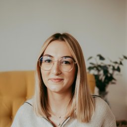 Poradnia psychoterapii MIND-TALK-THERAPY - Portret uśmiechniętej blondynki w okularach, siedzącej na żółtym fotelu. Za nią widoczna roślina doniczkowa i jasna ściana. Neutralny wyraz twarzy.