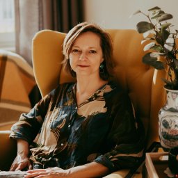 Poradnia psychoterapii MIND-TALK-THERAPY - Portret kobiety z krótkimi włosami siedzącej w żółtym fotelu. W tle widoczny jest pokój z dekoracjami i rośliną w wazonie. Naturalne światło.