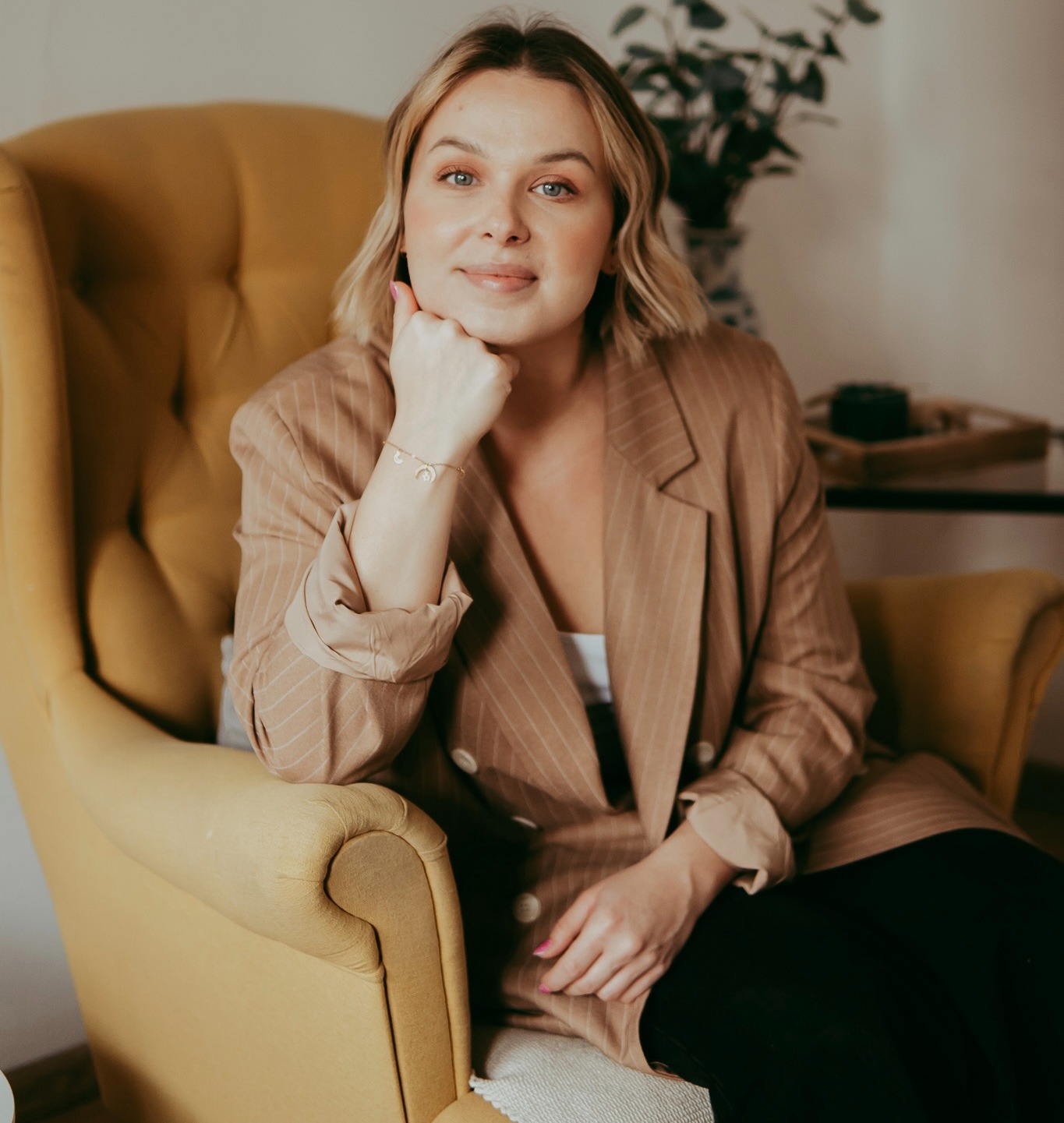 Portret kobiety z blond włosami w beżowym żakiecie, siedzącej w żółtym fotelu na tle jasnego wnętrza. Dłoń podparta pod brodą, subtelny uśmiech.