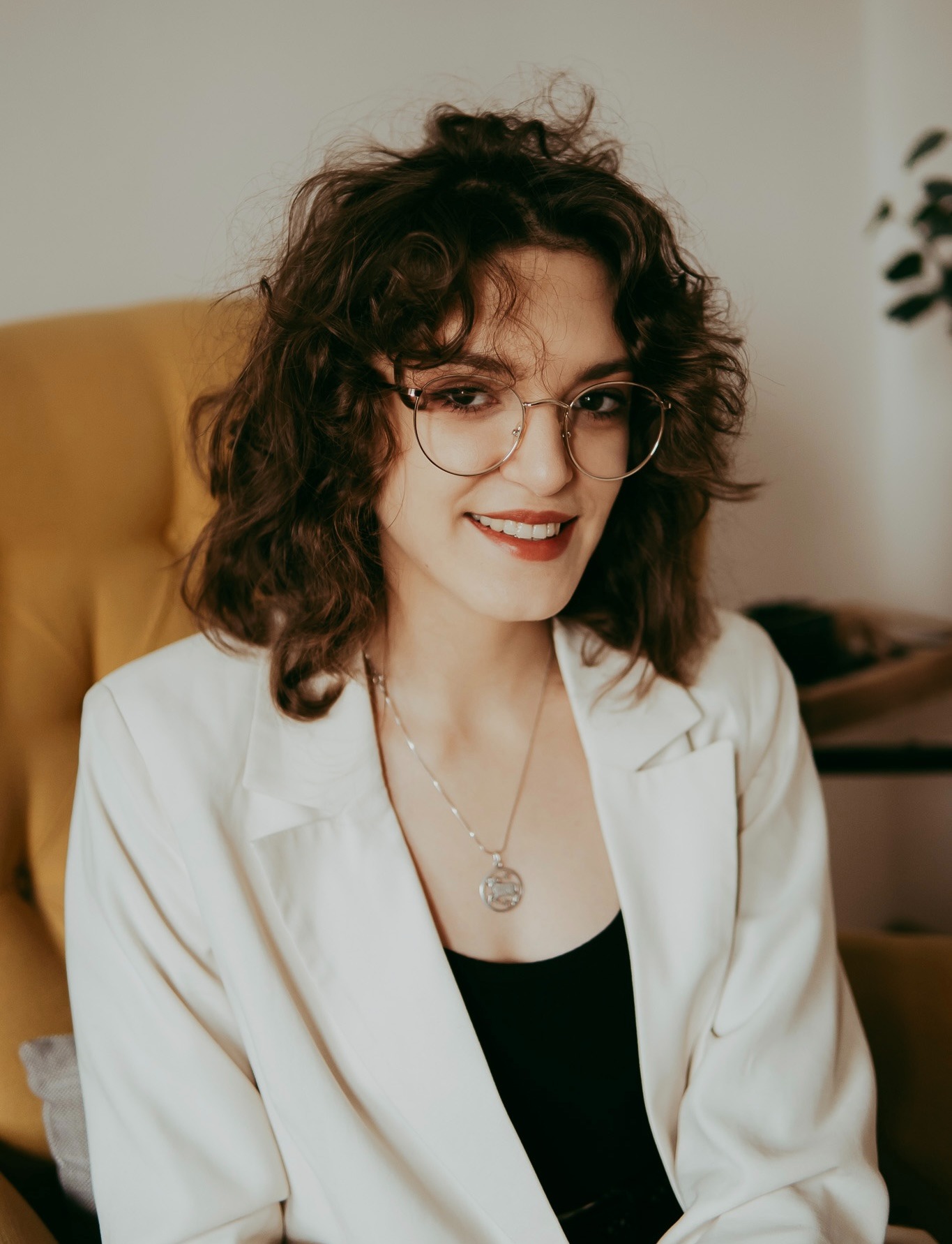 Uśmiechnięta kobieta w okularach i białej marynarce siedzi na żółtym fotelu, patrząc w obiektyw. W tle widoczny fragment rośliny doniczkowej i jasna ściana.