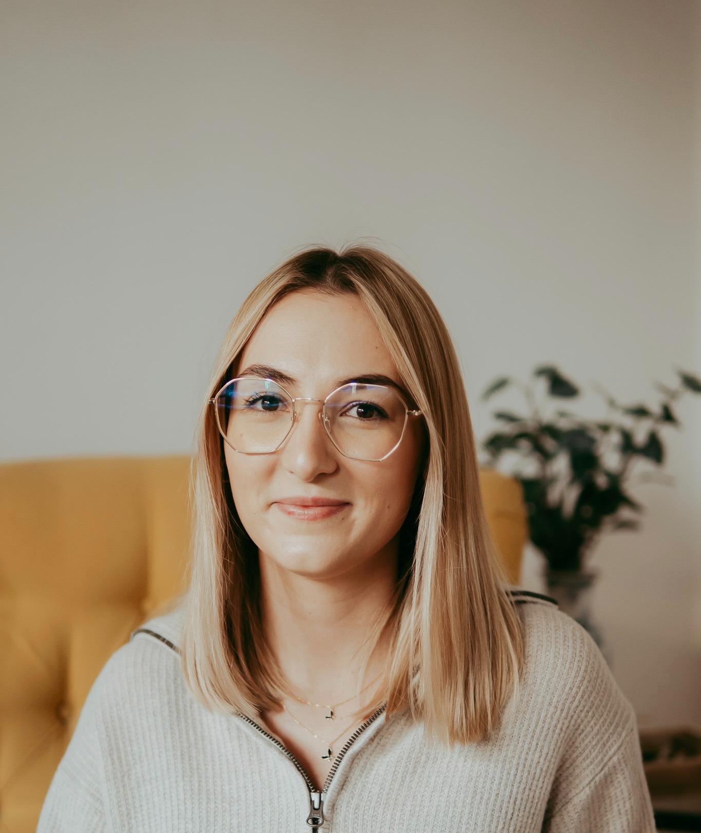 Portret uśmiechniętej blondynki w okularach, siedzącej na żółtym fotelu. Za nią widoczna roślina doniczkowa i jasna ściana. Neutralny wyraz twarzy.