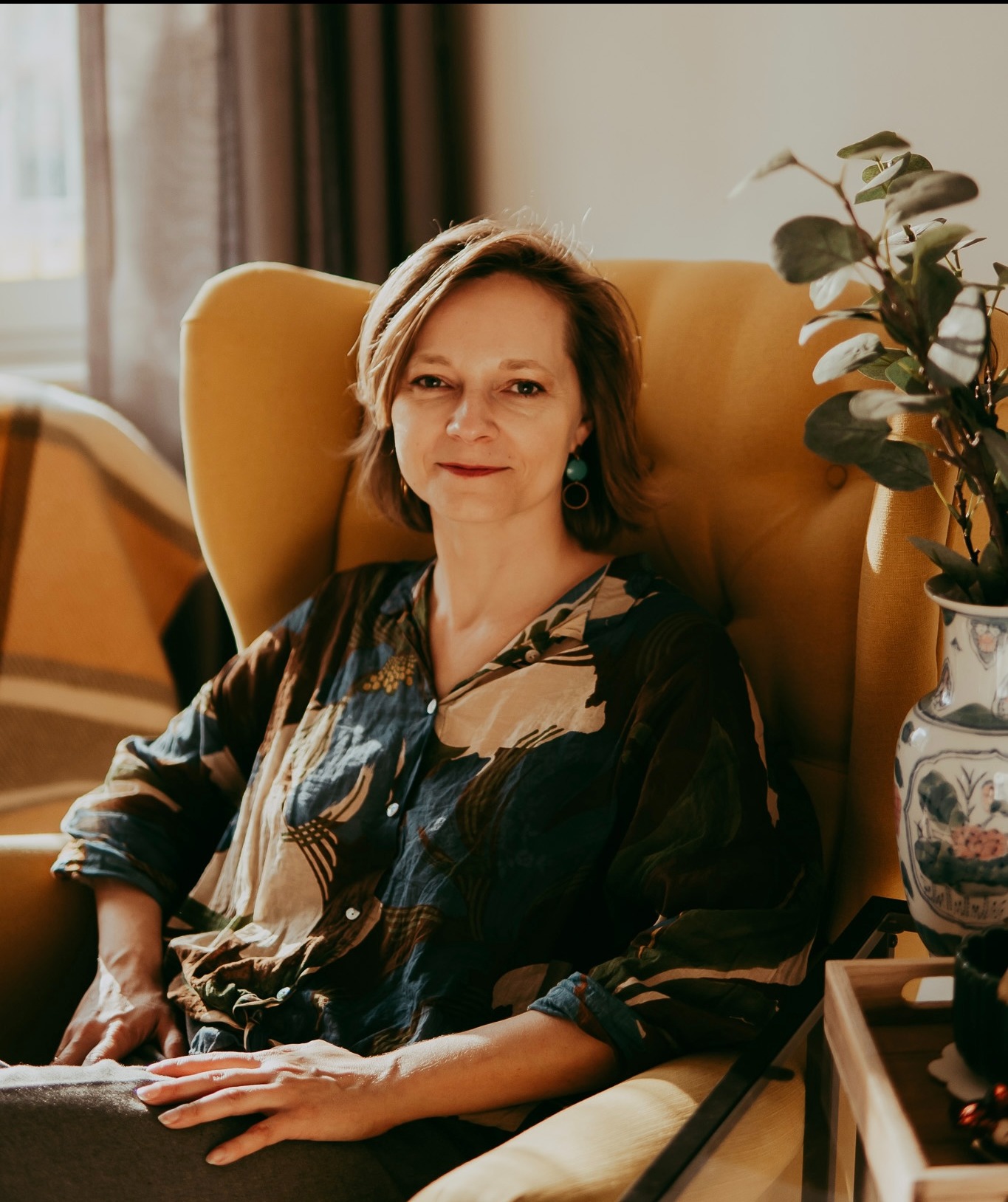 Portret kobiety z krótkimi włosami siedzącej w żółtym fotelu. W tle widoczny jest pokój z dekoracjami i rośliną w wazonie. Naturalne światło.