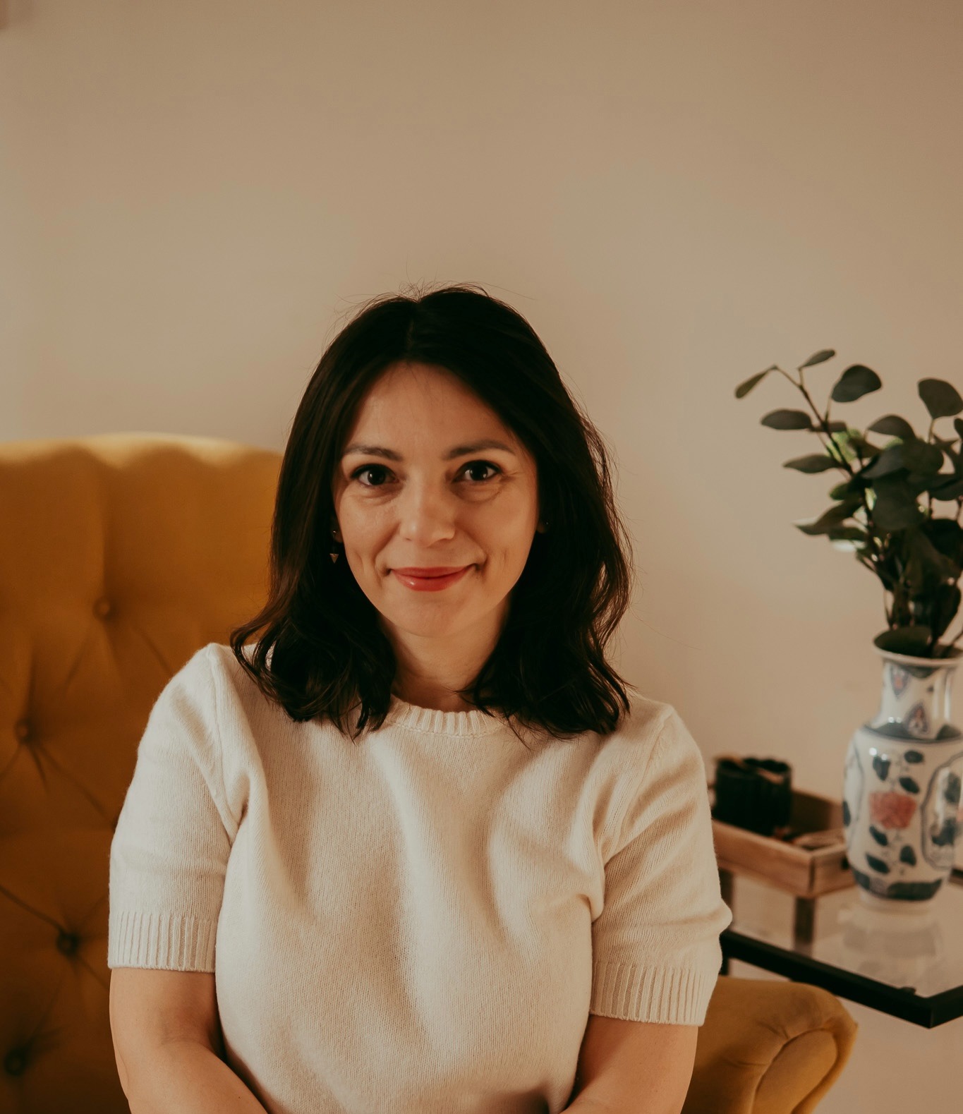 Uśmiechnięta kobieta w swetrze ecru siedzi na musztardowym fotelu. W tle wazon z gałązkami na szklanym stoliku. Neutralne tło, ciepłe światło.