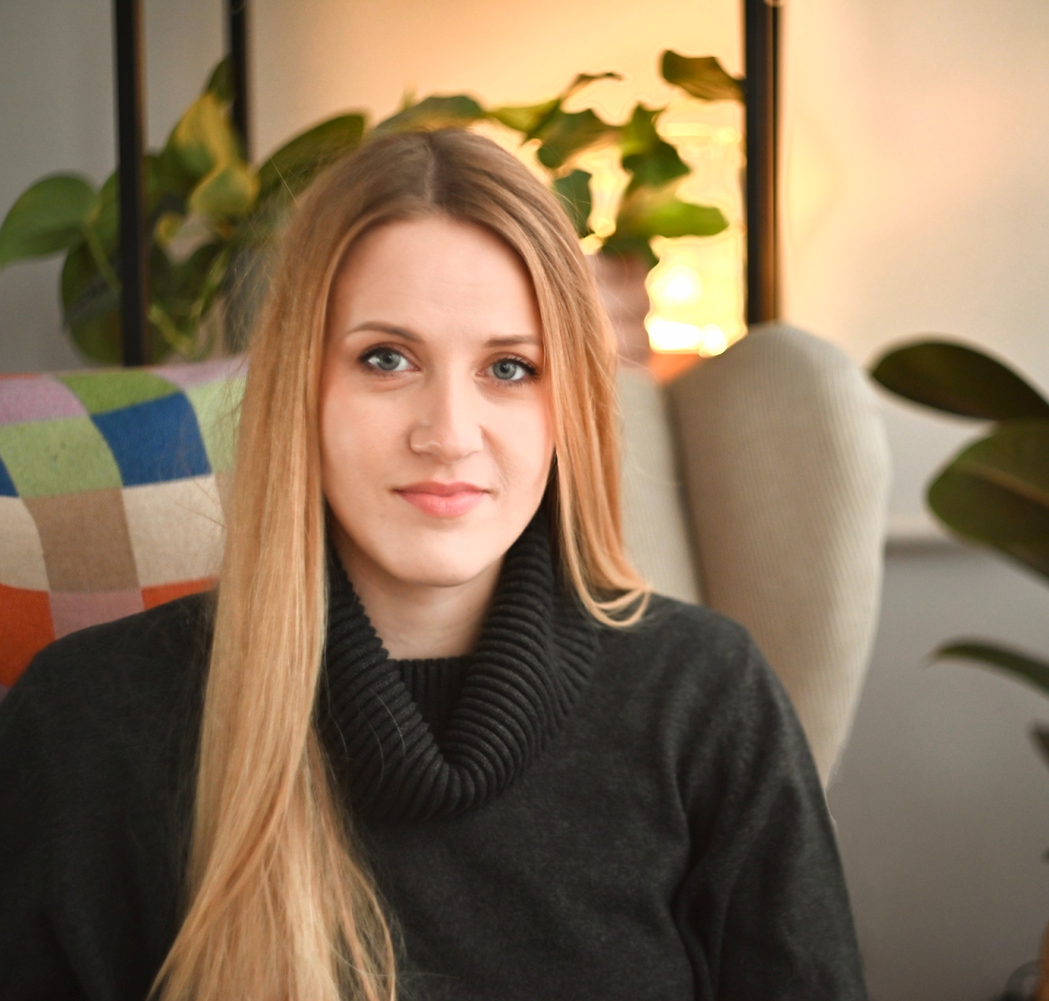 Portret kobiety o jasnych włosach i niebieskich oczach, siedzącej w fotelu na tle domowych roślin i kolorowej poduszki, w swetrze z golfem. Ciepłe, naturalne światło.