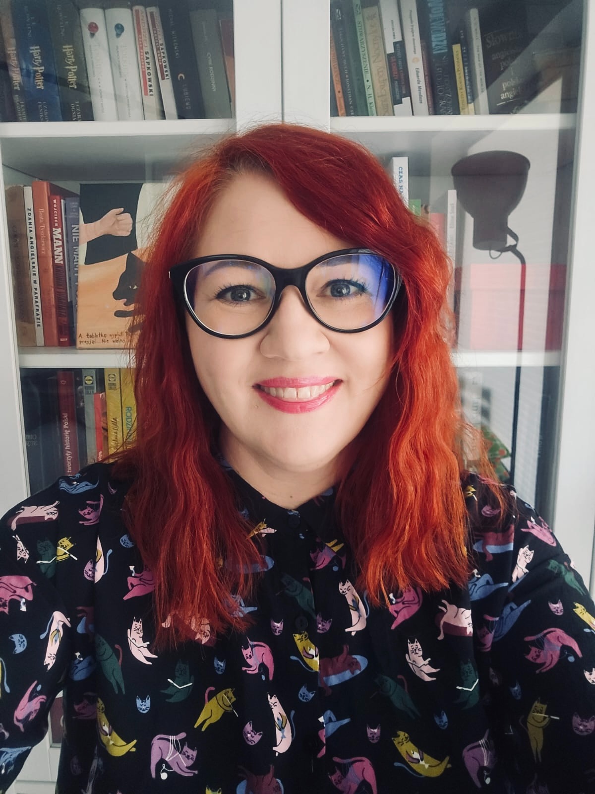 Portret uśmiechniętej kobiety z rudymi włosami i okularach na tle biblioteczki. Ubrana w koszulę w kolorowe koty. Ciepłe oświetlenie, bliski kadr.