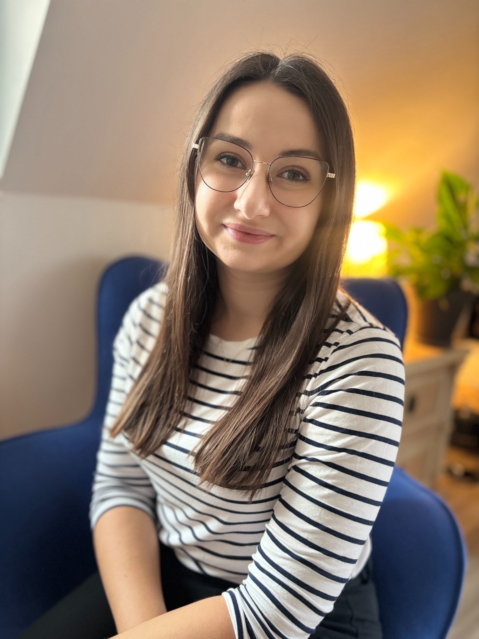 Portret uśmiechniętej kobiety w okularach, siedzącej na niebieskim fotelu. W tle widoczna lampa i roślina doniczkowa, tworzące przytulną atmosferę.