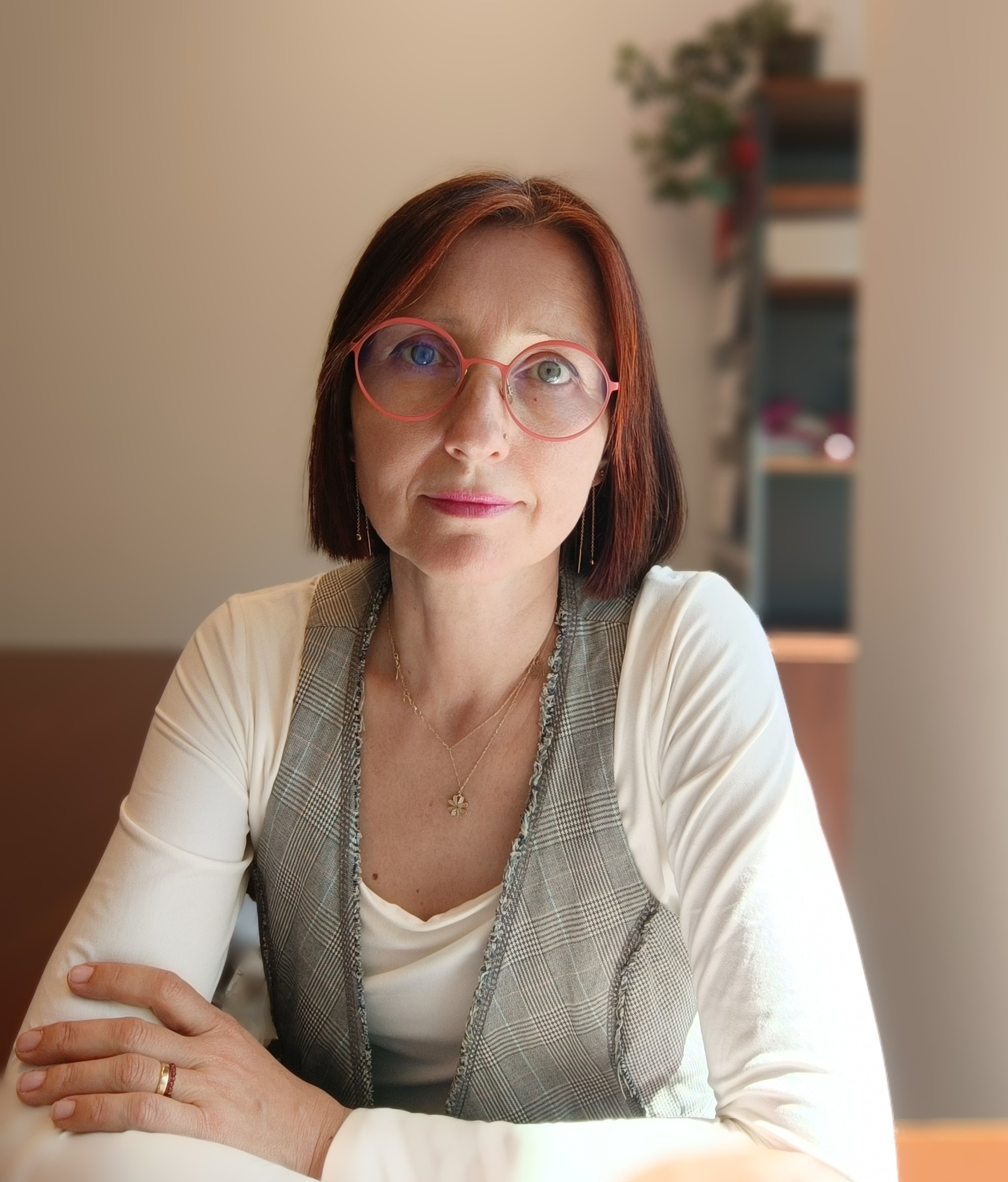 Portret kobiety w okularach z rudymi refleksami, ubrana w białą bluzkę i szarą kamizelkę w kratę, siedzącej za biurkiem. W tle rozmyte półki z książkami.