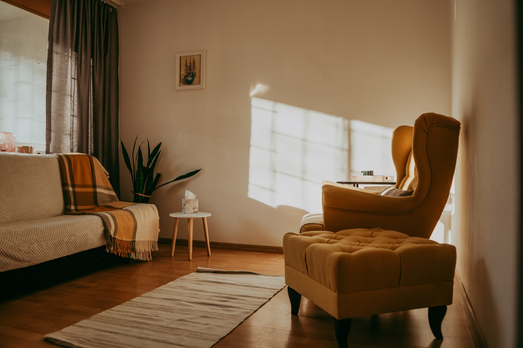 Jasny pokój z żółtym fotelem uszak, sofą z pledem, rośliną doniczkową i dywanem. W tle ściana z obrazkiem i promieniami słońca. Przytulne wnętrze.