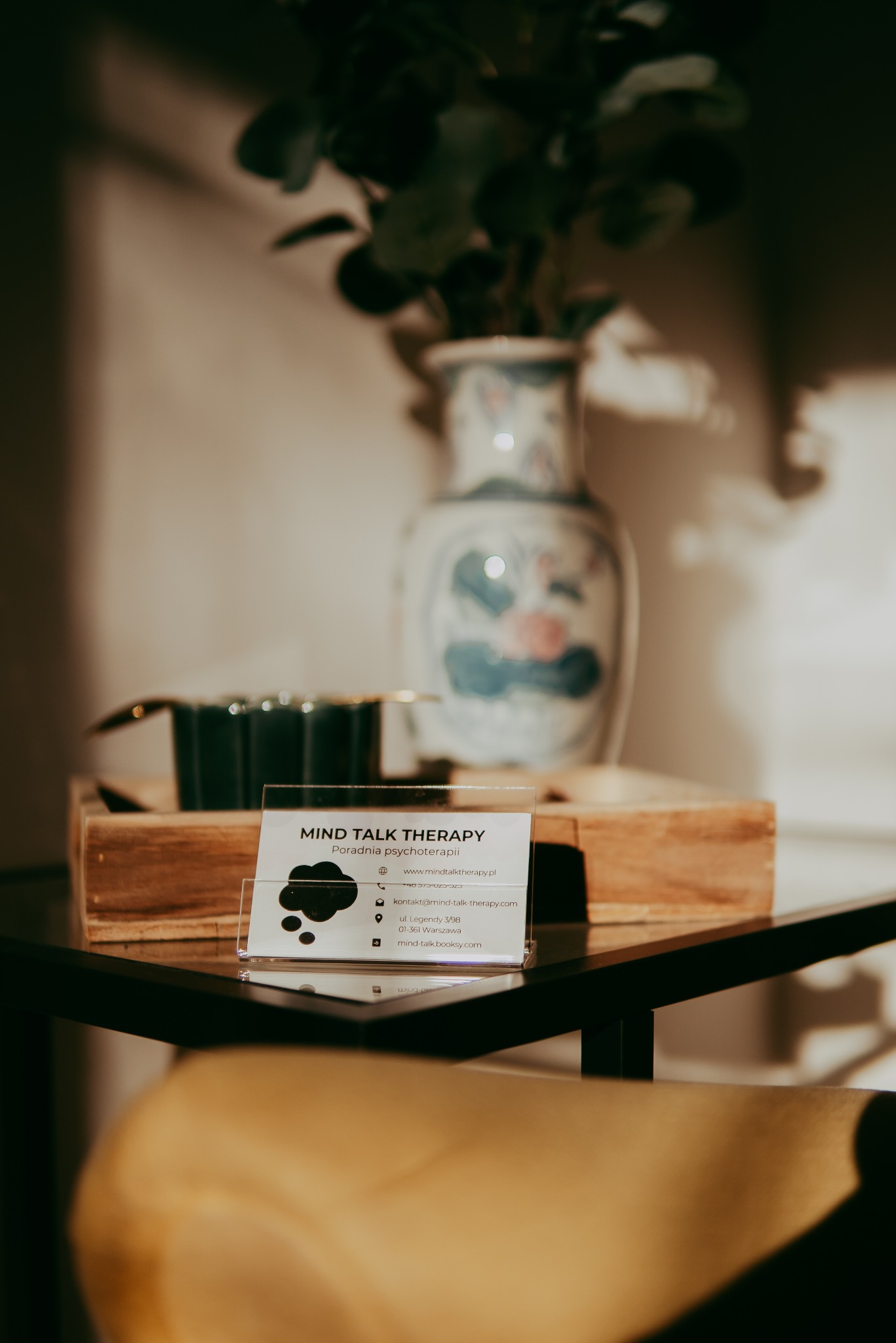 Wizytówka poradni psychoterapii 'Mind Talk Therapy' na stoliku, z dekoracyjnym wazonem w tle. Ciepłe oświetlenie tworzy przytulną atmosferę gabinetu.