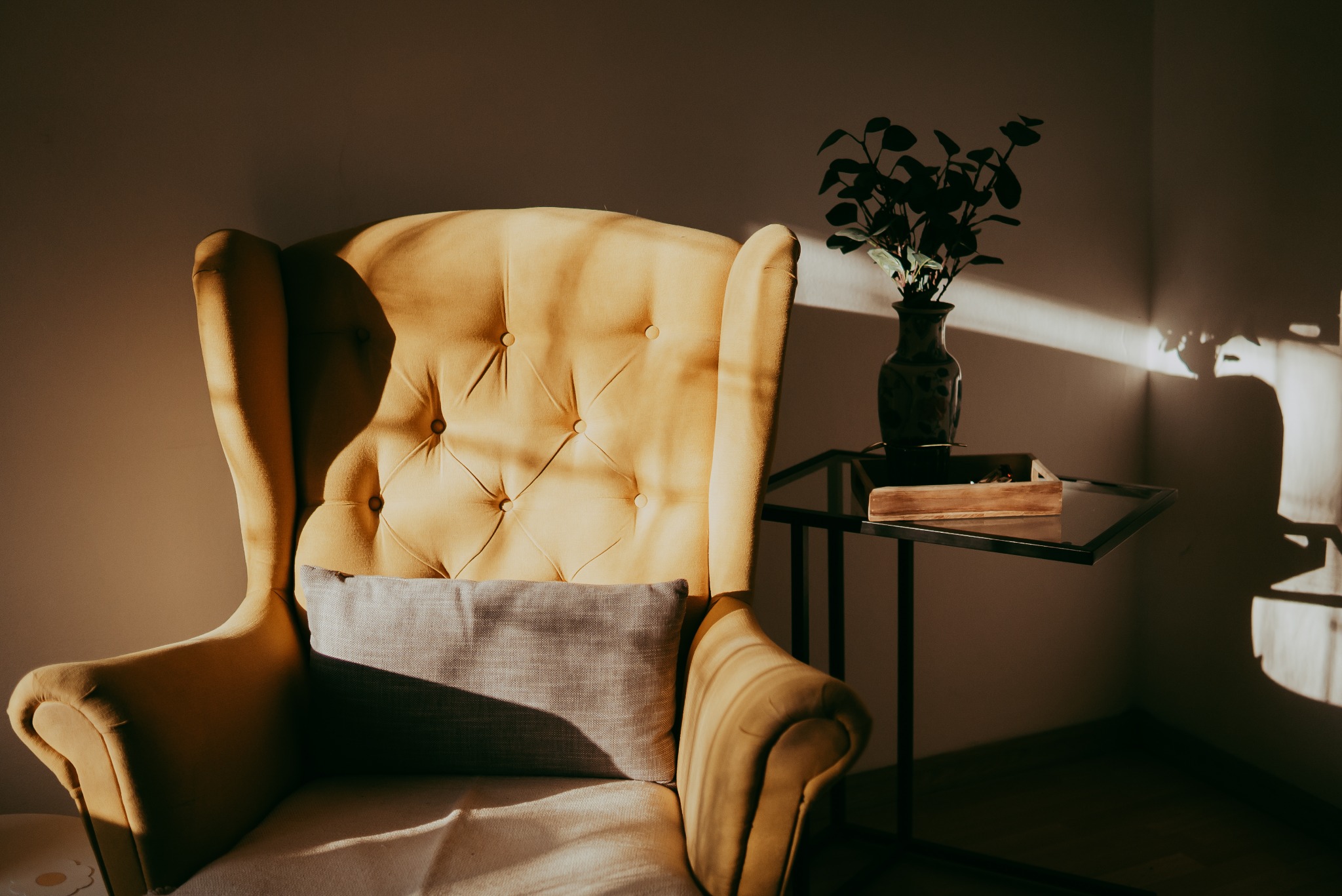 Żółty fotel typu uszak z poduszką w pokoju, obok stolik z wazonem. Ciepłe światło słoneczne tworzy nastrojowe cienie na ścianie. Komfortowe wnętrze.
