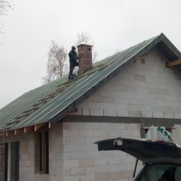 Andrzej Sulik - Budowa domu z białych bloczków: murarz na dachu układa łatki pod pokrycie, komin z cegły, otwarty bagażnik auta w tle, drzewa bez liści w zimowej scenerii.