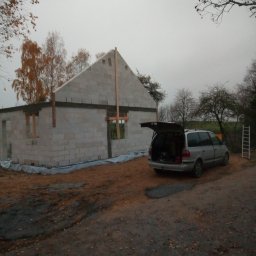 Andrzej Sulik - Surowy stan domu z szarego bloczka, drewniane stemple podtrzymujące konstrukcję. Na podwórku stoi srebrny samochód z otwartym bagażnikiem, jesienne drzewa w tle.