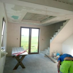 Andrzej Sulik - Wnętrze w trakcie prac wykończeniowych: gładzie na ścianach i suficie, widoczne płyty gipsowe i instalacje. Stolik roboczy i osoba w jaskrawych spodniach w pomieszczeniu.