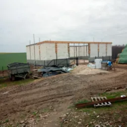 Surowy stan budynku z betonu komórkowego w trakcie budowy, widoczne zbrojenia, teren budowy z piaskiem, mieszarką i przyczepą. Pochmurne niebo w tle.