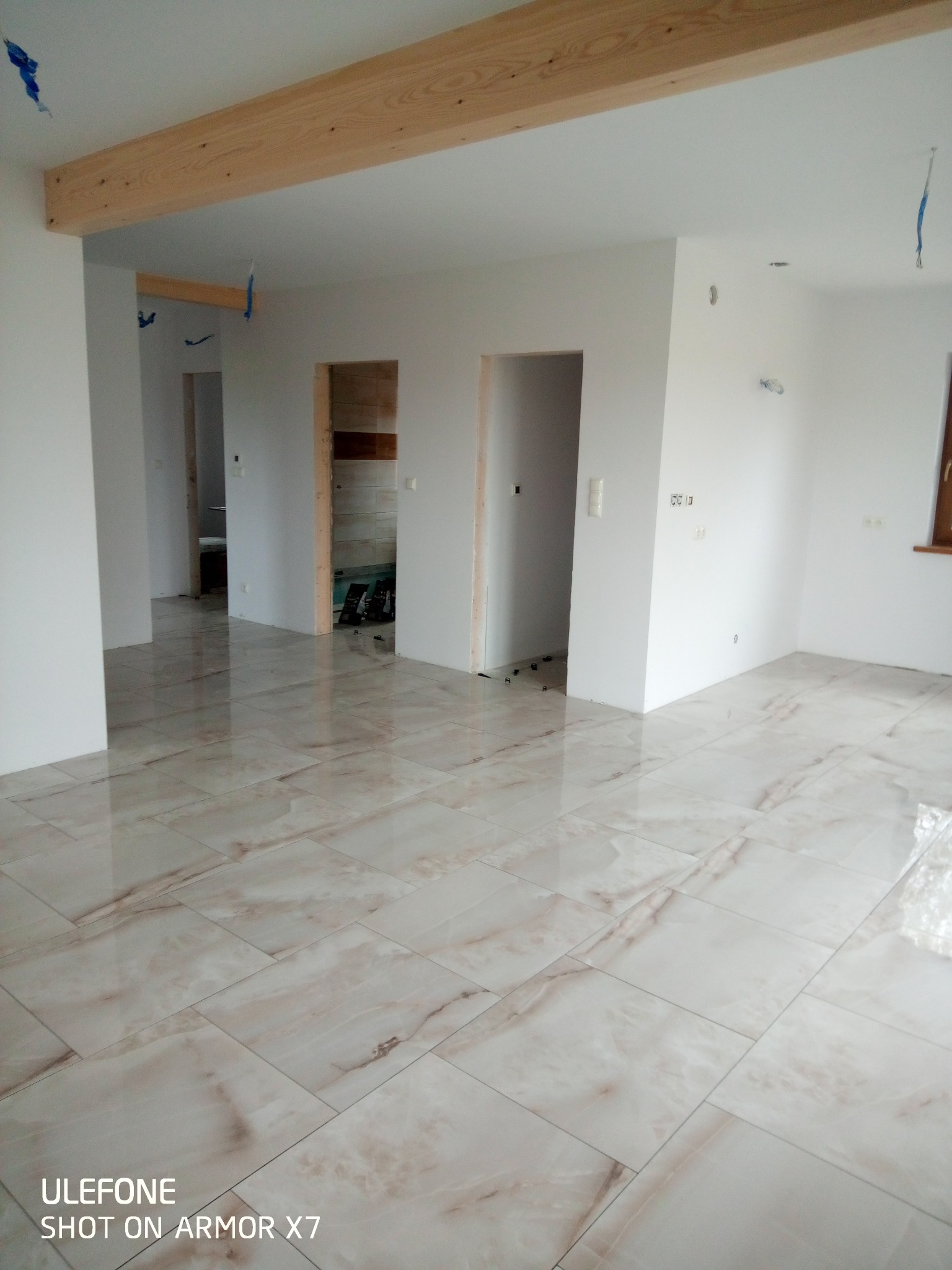 Jasne płytki podłogowe z marmurkowym wzorem w przestronnym, jasnym wnętrzu z surowymi belkami stropowymi i otworami drzwiowymi.