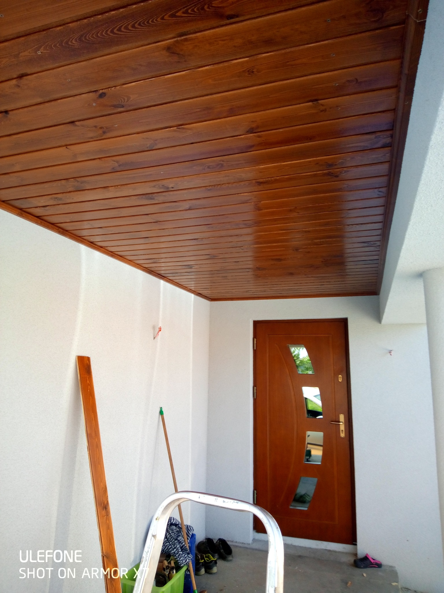 Drewniany sufit z desek w kolorze miodowym, białe ściany i brązowe drzwi wejściowe z szybkami. Pod ścianą oparta drabina i listwa. Widok z dołu.