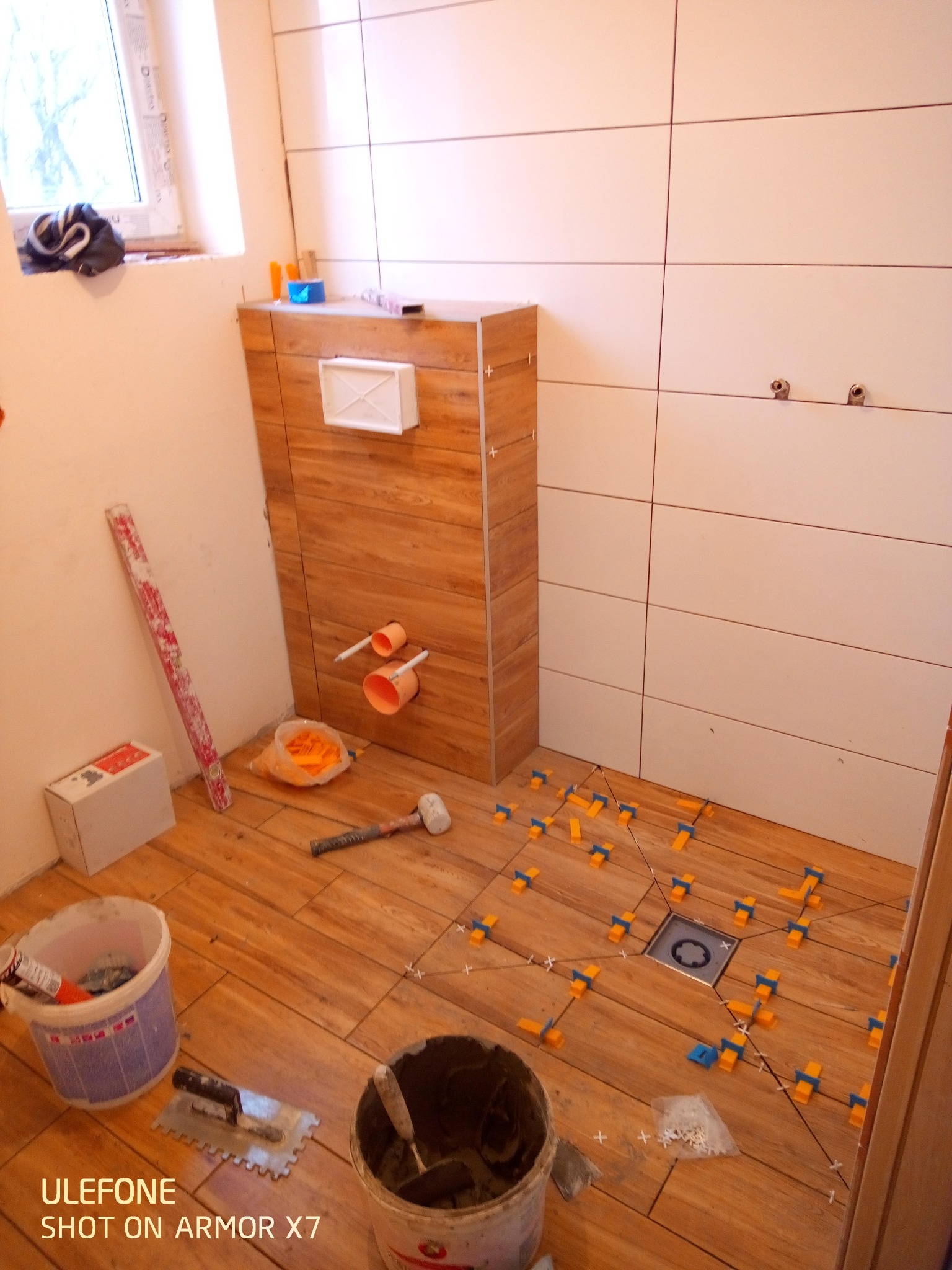 Układanie płytek imitujących drewno na podłodze w łazience. Widoczne narzędzia, poziomica, wiadro z klejem i krzyżyki dystansowe. Częściowo ułożone płytki na ścianie.