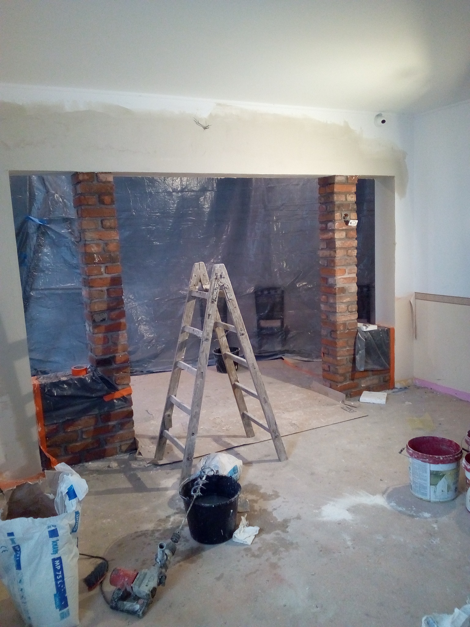 Wnętrze w trakcie remontu: widoczne ceglane filary, drabina, worki z zaprawą, wiadro i mieszadło. Folia ochronna zabezpiecza dalszą część pomieszczenia.