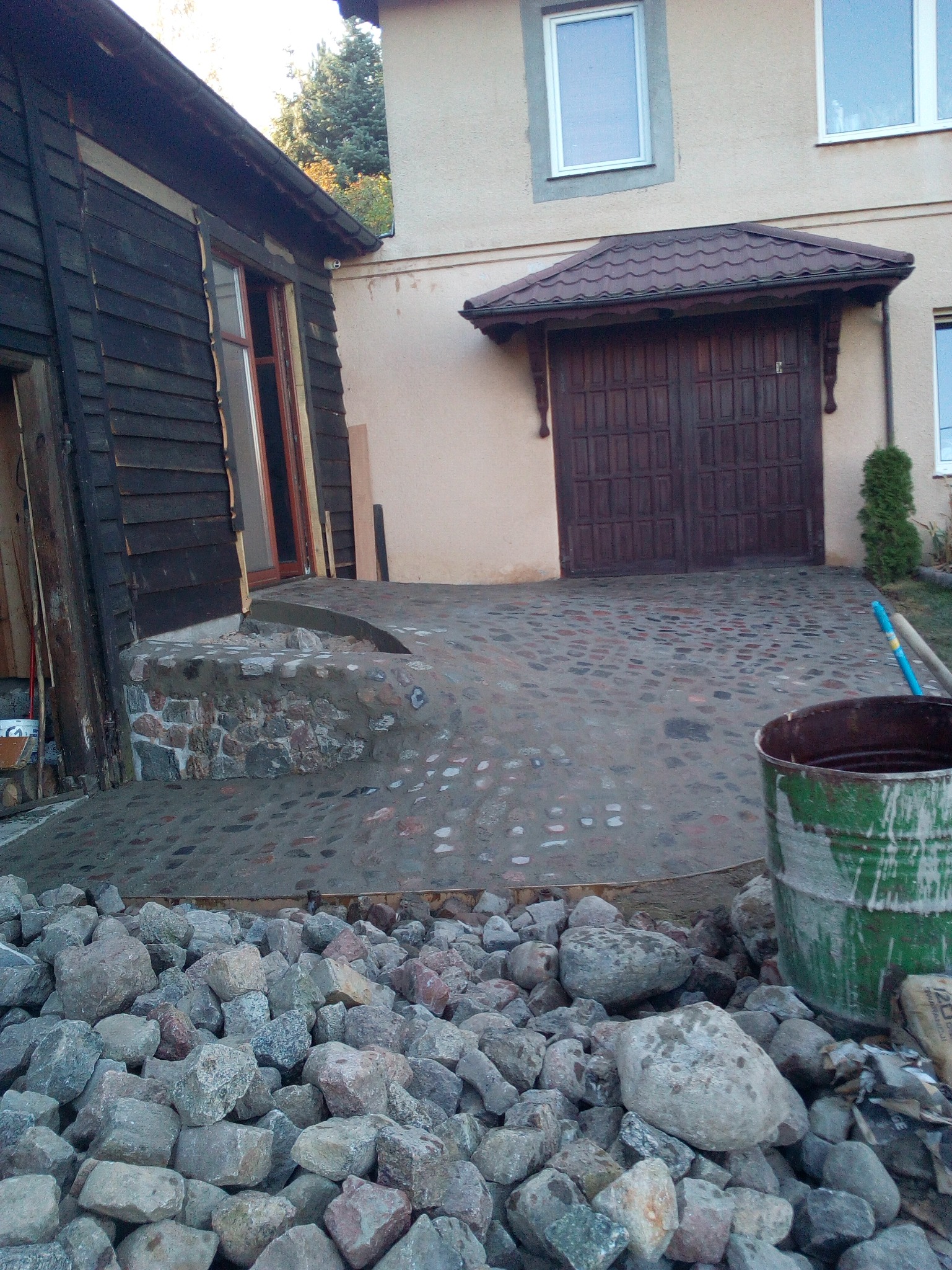 Ułożona kostka brukowa na podjeździe z kamiennymi schodami i starym budynkiem w tle. Widoczne narzędzia i materiały budowlane. Charakterystyczny wzór kostki.