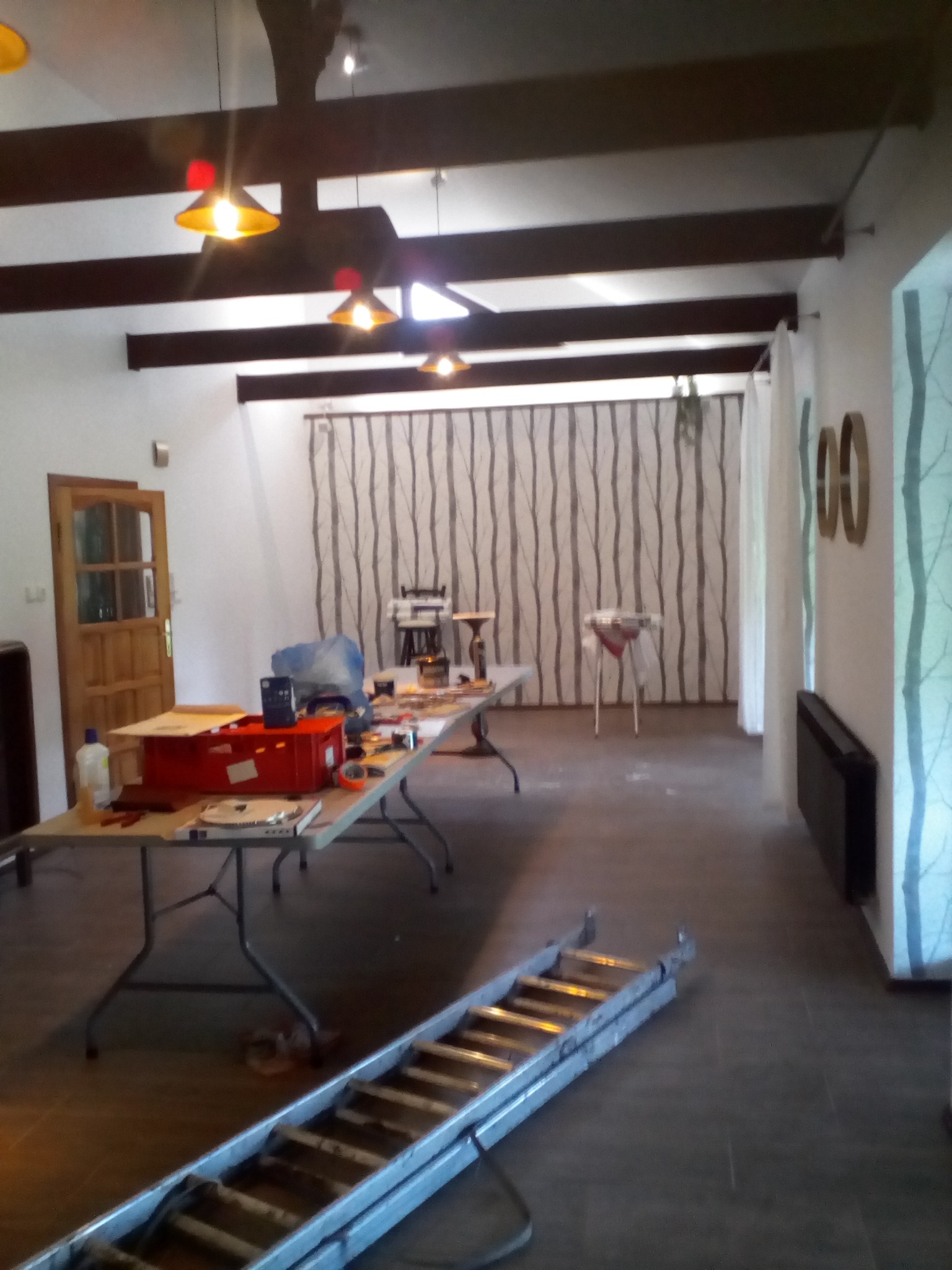 Przygotowania do prac wykończeniowych w Biskupcu. Rozłożona drabina, stół z narzędziami i materiałami w przestronnym wnętrzu z drewnianymi belkami stropowymi.