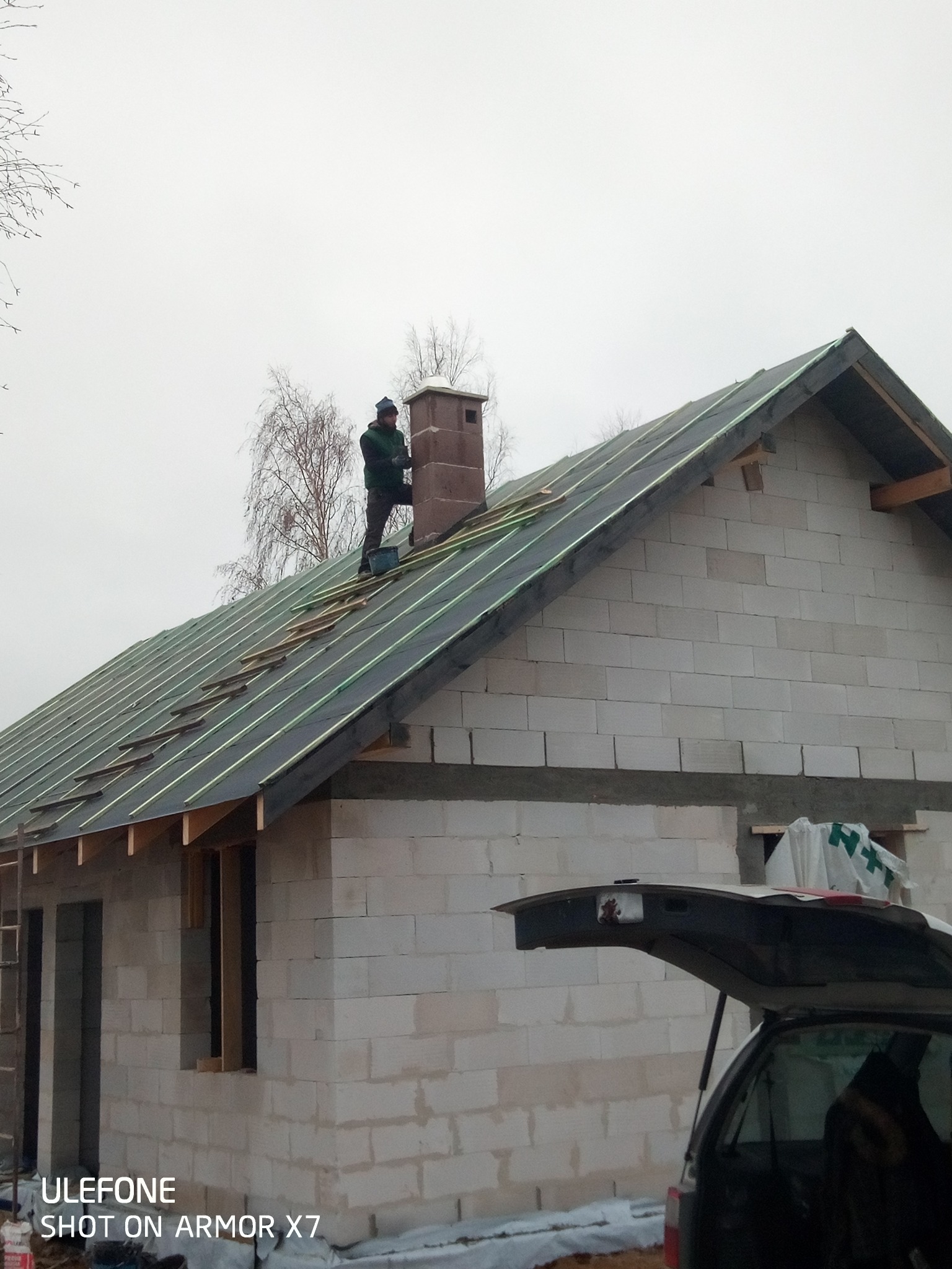 Budowa domu z białych bloczków: murarz na dachu układa łatki pod pokrycie, komin z cegły, otwarty bagażnik auta w tle, drzewa bez liści w zimowej scenerii.