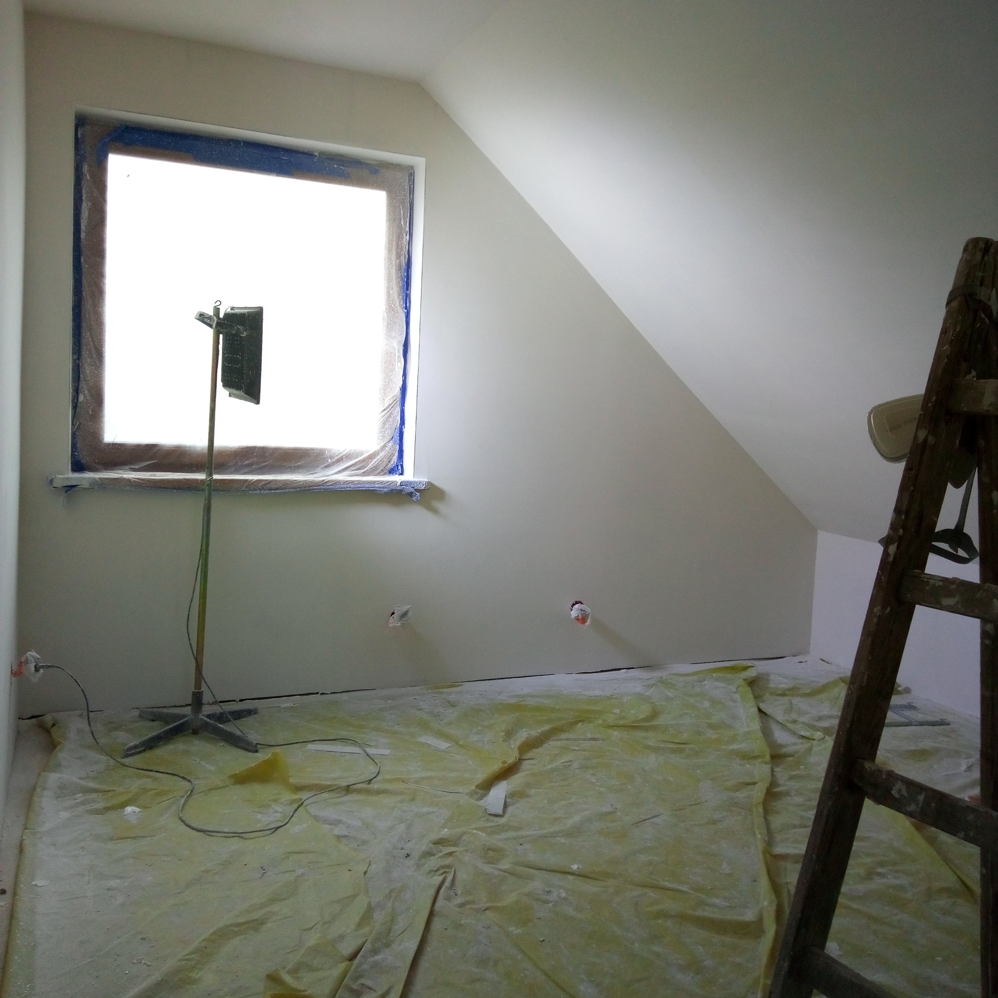 Pokój na poddaszu po gładzi, zabezpieczony folią, z oknem i stojącą lampą halogenową. Widoczna drabina malarska.
