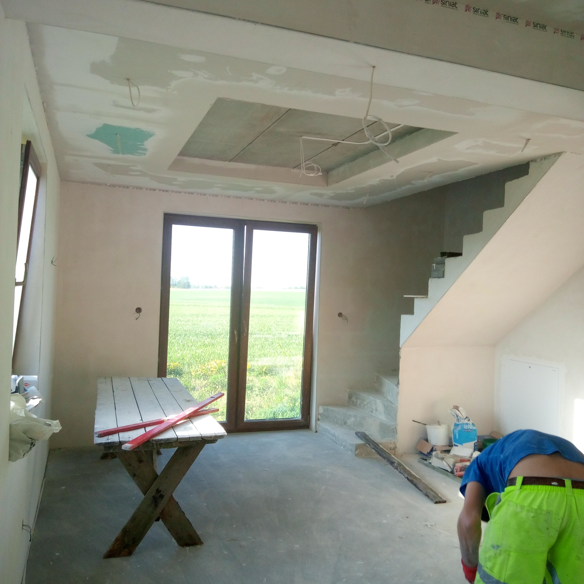 Wnętrze w trakcie prac wykończeniowych: gładzie na ścianach i suficie, widoczne płyty gipsowe i instalacje. Stolik roboczy i osoba w jaskrawych spodniach w pomieszczeniu.