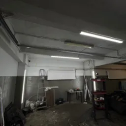 Wnętrze warsztatu w Rzeszowie z zamontowanym oświetleniem LED na suficie. Widoczne przewody i elementy instalacji. Biało-szare ściany, drabina i narzędzia.