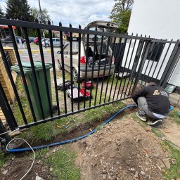 Voltarius - Instalacja okablowania do bramy automatycznej. Widoczny pracownik w trakcie pracy, narzędzia, skrzynka narzędziowa i częściowo otwarty bagażnik samochodu.