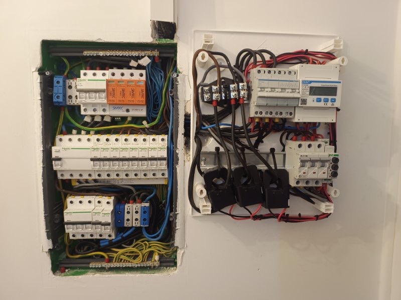 Dwie rozdzielnice elektryczne w ścianie: jedna zielona z bezpiecznikami, druga biała od fotowoltaiki, z widocznymi kablami i elementami zabezpieczeń.