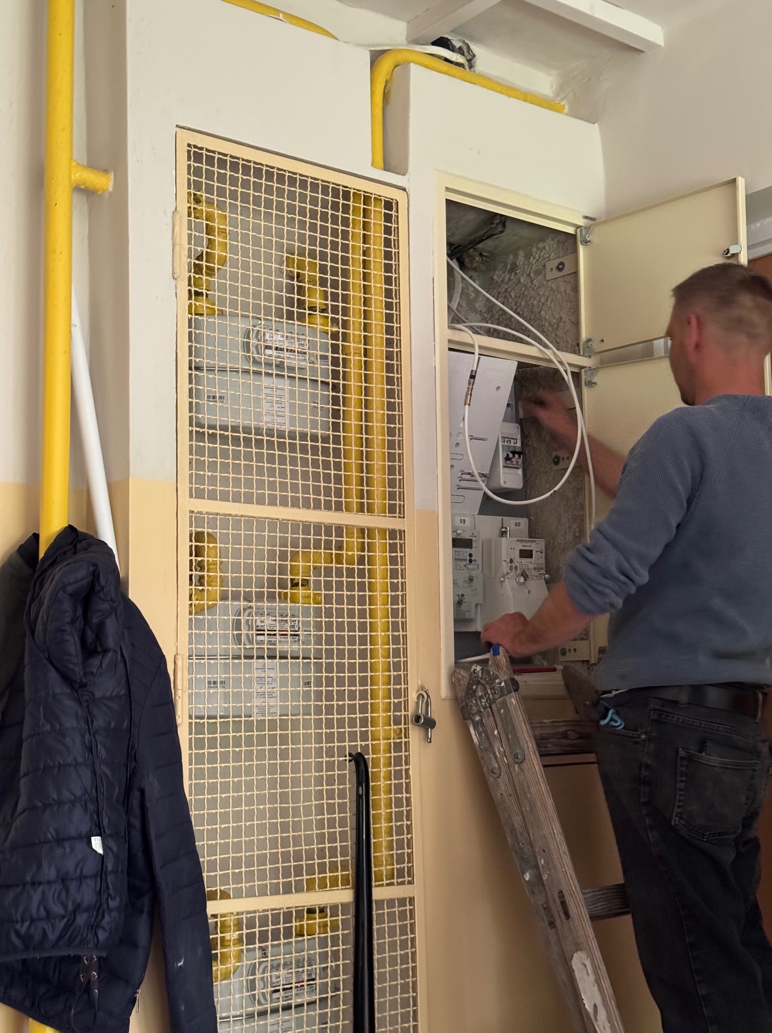 Elektryk na drabinie, w trakcie prac przy skrzynce z licznikami w żółtej obudowie z kratą. Widoczne przewody i elementy instalacji.