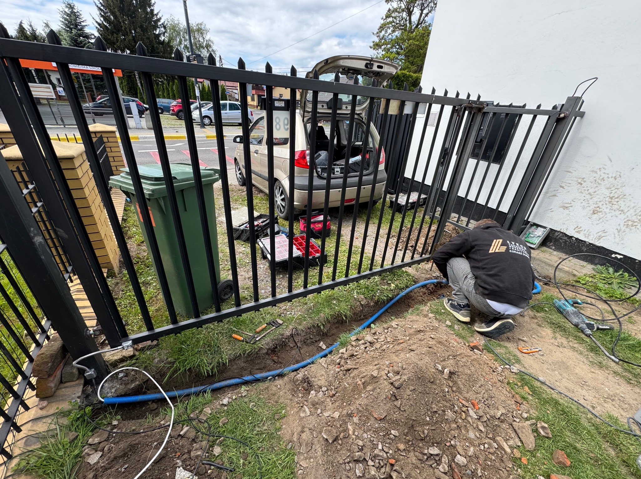 Instalacja okablowania do bramy automatycznej. Widoczny pracownik w trakcie pracy, narzędzia, skrzynka narzędziowa i częściowo otwarty bagażnik samochodu.