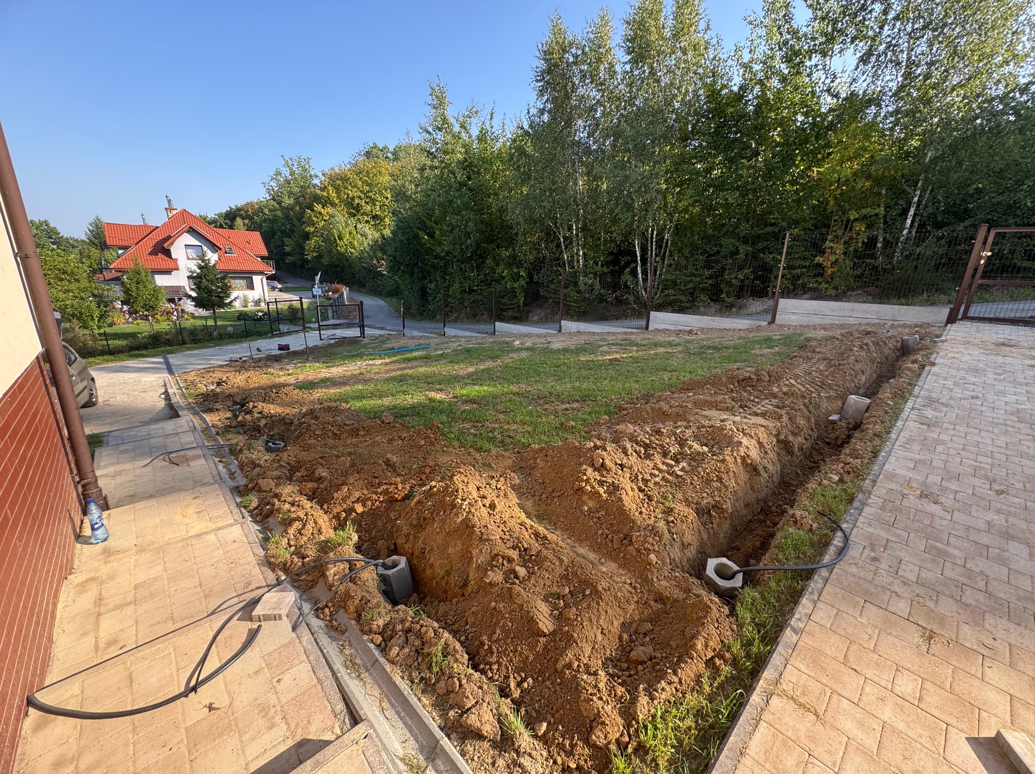 Wykopane rowy w ziemi pod instalację, widoczne kable i betonowe elementy. W tle dom z czerwoną dachówką i drzewa. Ujęcie z góry, słoneczny dzień.