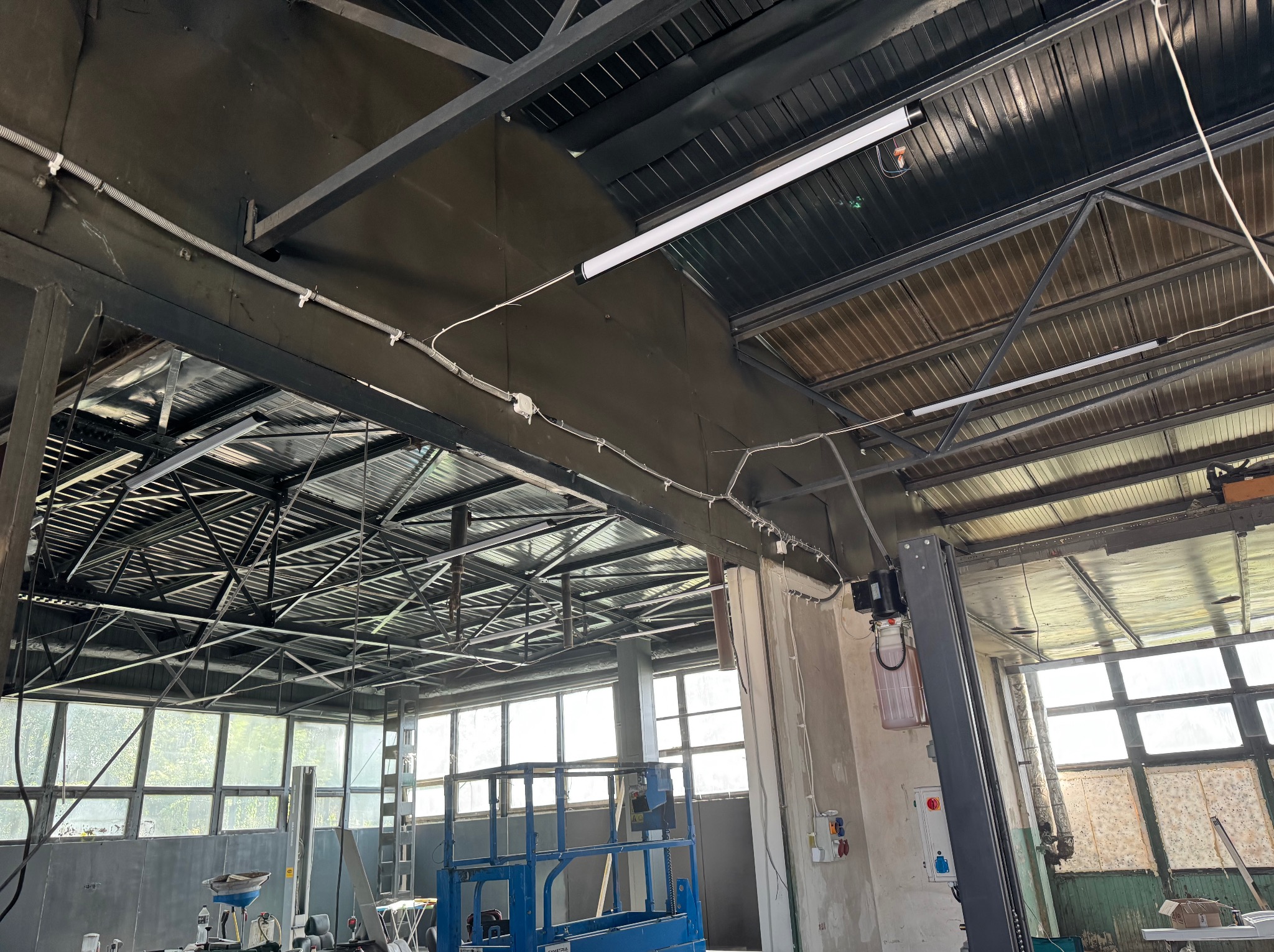 Wnętrze hali warsztatowej z widoczną instalacją elektryczną pod sufitem, oświetleniem LED i konstrukcją stalową. Widoczny podnośnik samochodowy i okna.
