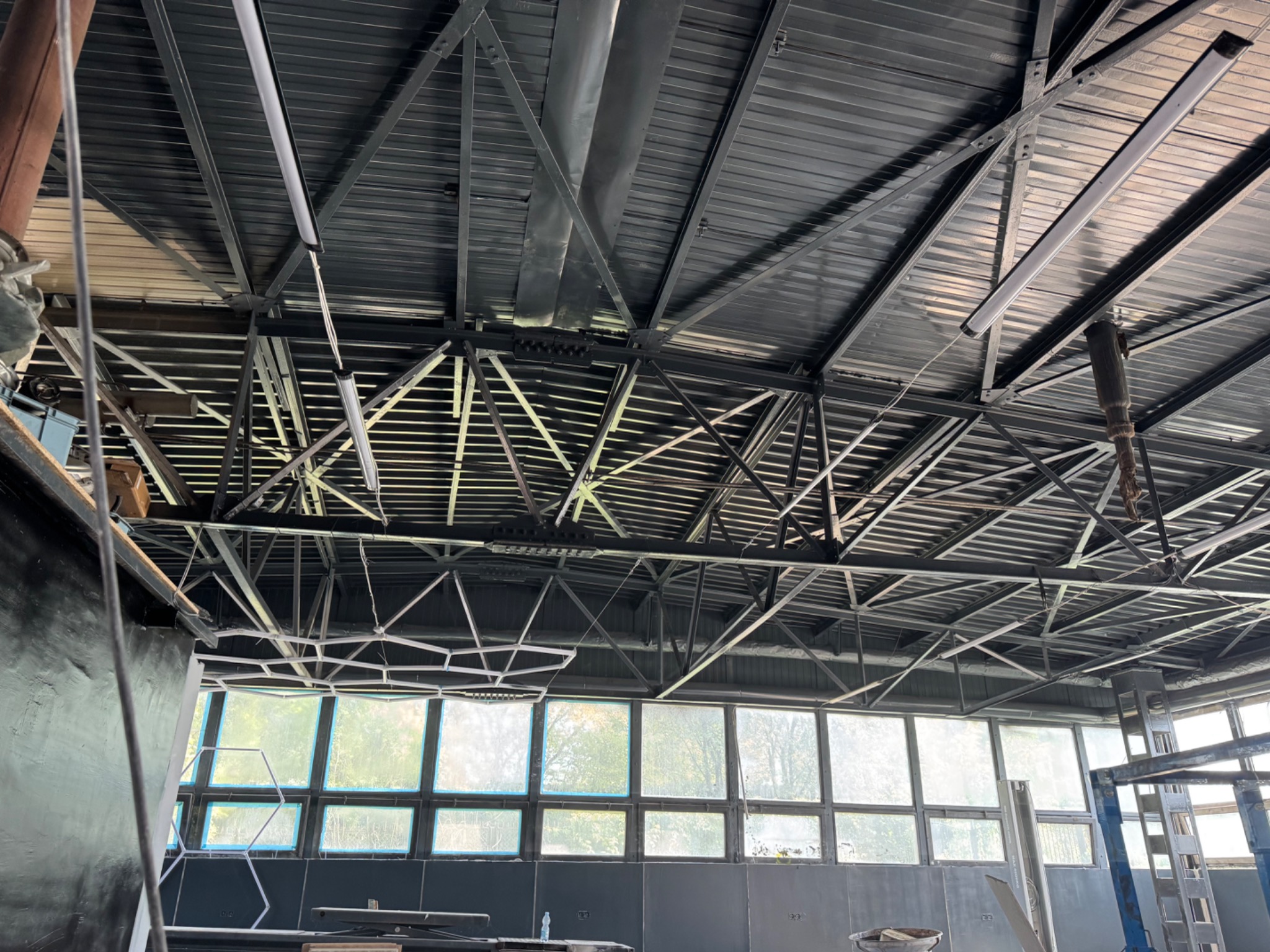 Metalowa konstrukcja dachu hali z oknami w tle. Widoczne rury wentylacyjne, oświetlenie liniowe i elementy konstrukcyjne. Surowy, industrialny charakter wnętrza.