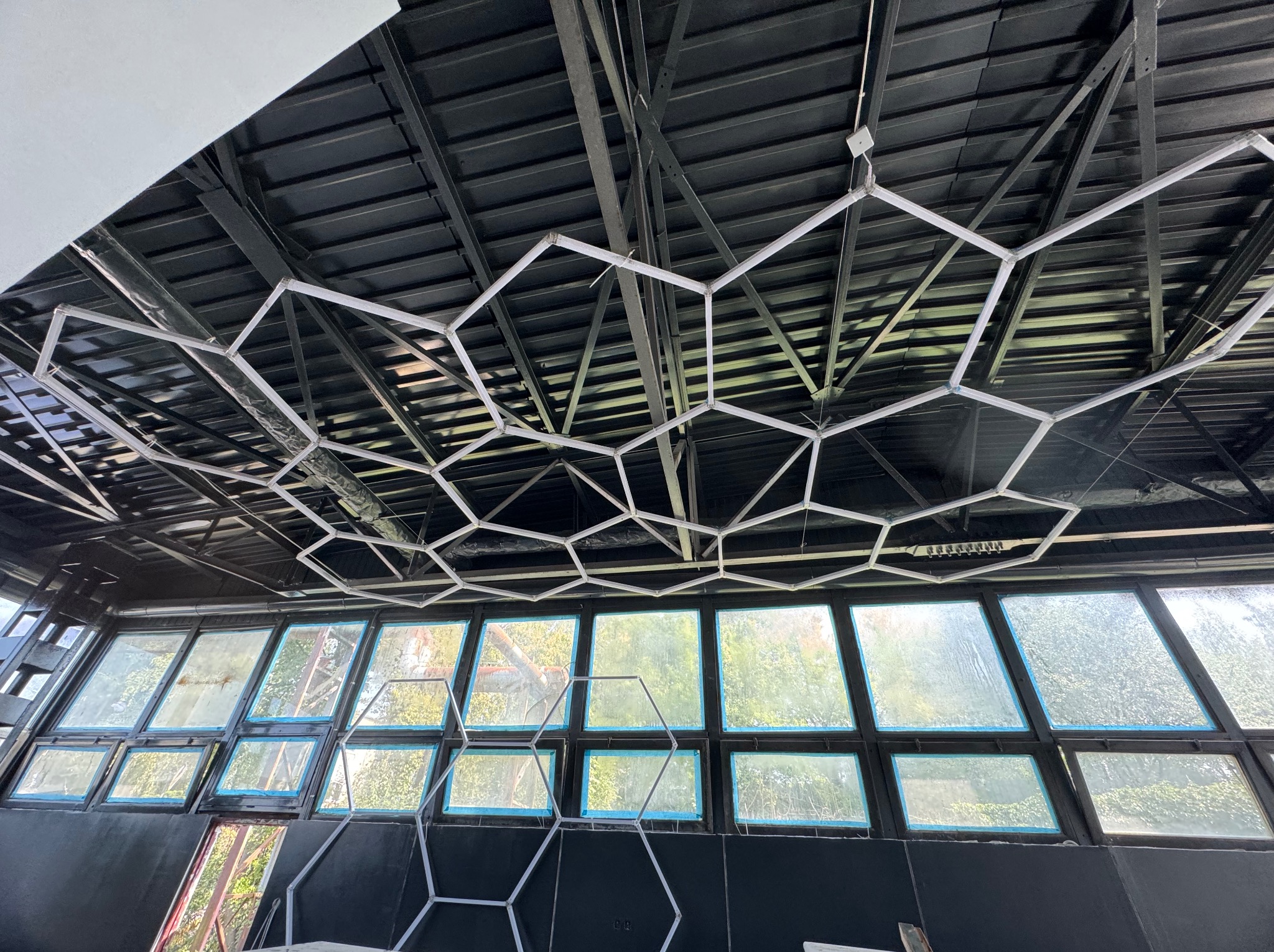 Nowoczesna instalacja oświetleniowa w kształcie plastra miodu, zawieszona pod czarnym, metalowym sufitem w przeszklonym pomieszczeniu z widokiem na zieleń.