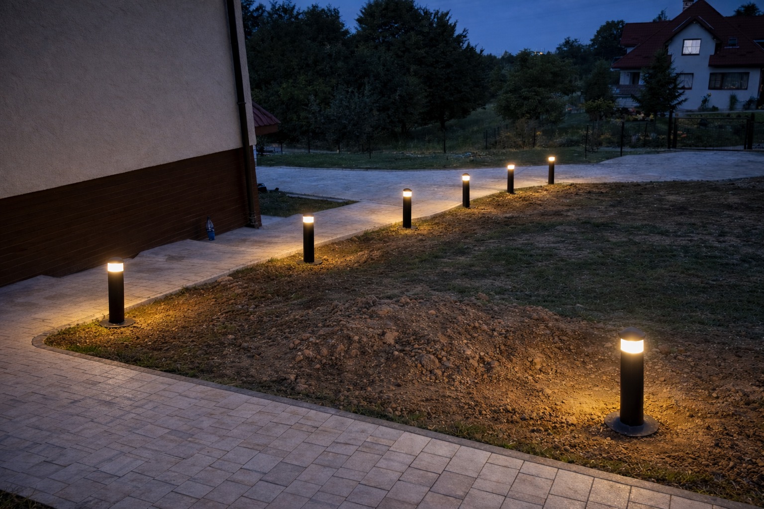 Ścieżka oświetlona szeregiem lamp ogrodowych w kształcie słupków, prowadząca do domu z czerwonym dachem, sfotografowana o zmierzchu.