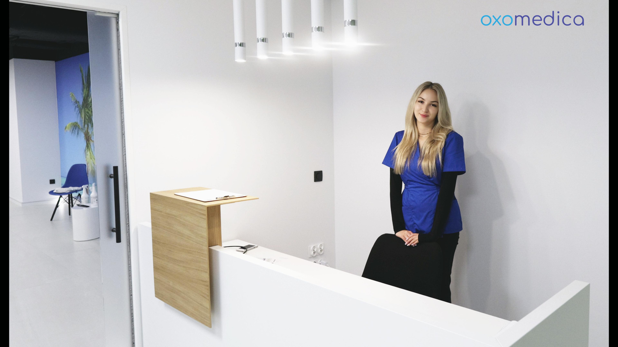 Jasne wnętrze kliniki Oxomedica: recepcja z uśmiechniętą blondynką w niebieskim uniformie, nowoczesne oświetlenie i minimalistyczny design.