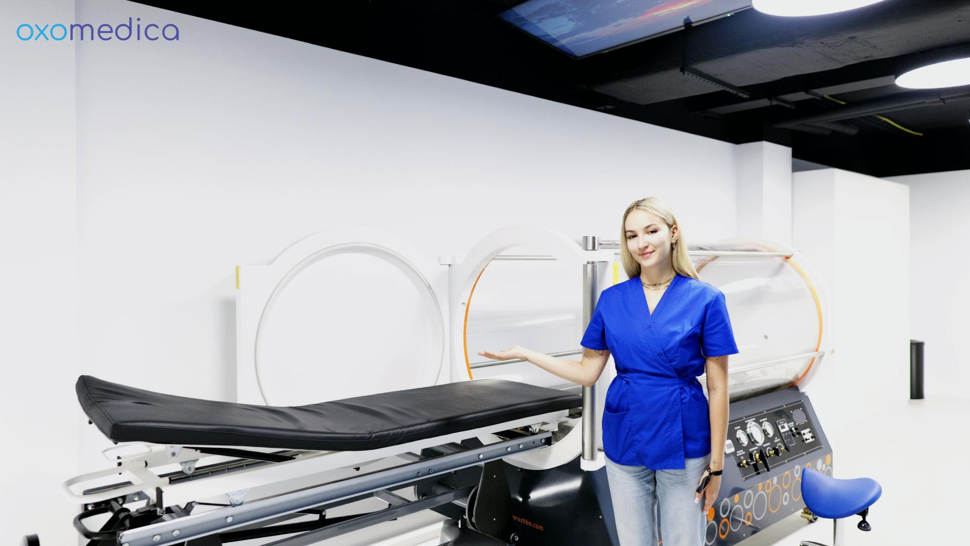 Kobieta w niebieskim uniformie prezentuje komorę hiperbaryczną w nowoczesnej klinice Oxomedica, z widocznym logo i oświetleniem sufitowym.