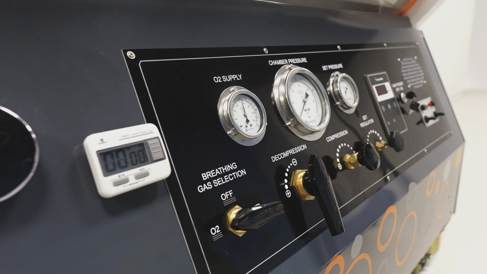 Panel sterowania komory hiperbarycznej z manometrami, przełącznikami i timerem. Widoczne napisy: O2 Supply, Chamber Pressure, Breathing Gas Selection.